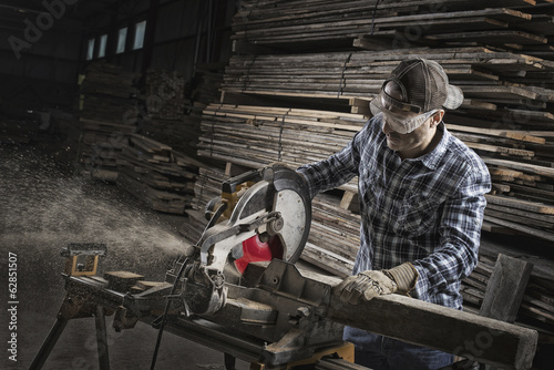 Foto: A reclaimed lumber workshop. A man in protective eye goggles using a circular saw to cut timber. A reclaimed lumber workshop. A man in protective eye goggles using a circular saw to cut timber.