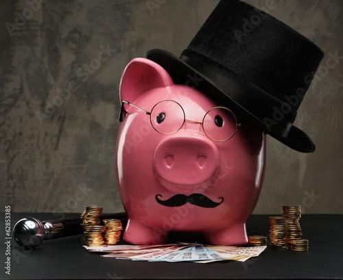 Photo: Piggybank in glasses and hat with pile of coins and banknotes Piggybank in glasses and hat with pile of coins and banknotes