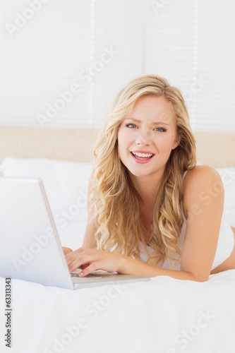 Photo: Smiling young blonde lying on her bed using laptop Smiling young blonde lying on her bed using laptop