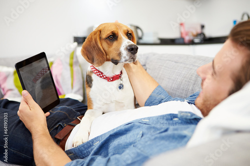 Photo: Young Man With Dog Sit On Sofa Using Digital Tablet Young Man With Dog Sit On Sofa Using Digital Tablet