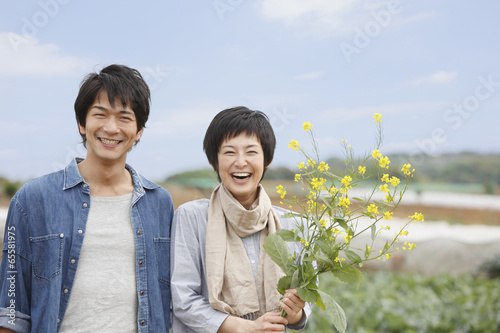 Photo: 畑の前で菜の花を持って立つ男女 畑の前で菜の花を持って立つ男女