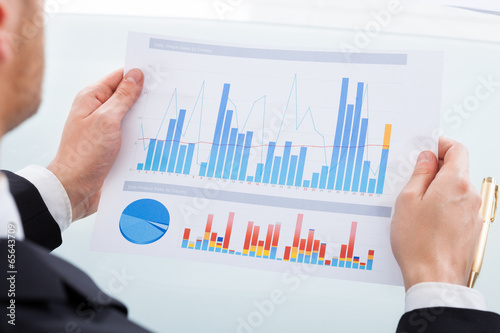 Photo: Businessman Comparing Graphs At Office Desk Businessman Comparing Graphs At Office Desk