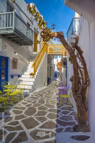  Streetview of Mykonos town with yellow chairs and tables and stairs, Greece