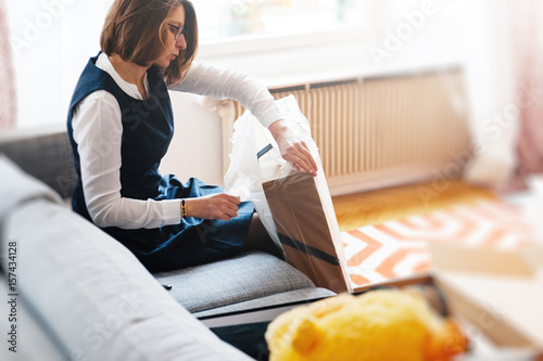 Plakat Młoda piękna kobieta na salonie sofa rozpakowuje unbox pudełko kartonowe zawierające nowe piękne ubrania
