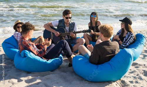 Plakat grupa przyjaciół z gitarą na imprezie na plaży - młodzi ludzie hipster na wakacje siedzi na torby fasoli i gra na gitarze w pobliżu morza