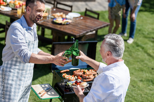 Zdjęcie XXL uśmiechnięci mężczyzna clinking butelki piwo podczas grilla outdoors