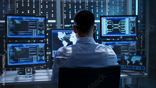 Фото: System Security Specialist Working at System Control Center. Room is Full of Screens Displaying Various Information. System Security Specialist Working at System Control Center. Room is Full of Screens Displaying Various Information.