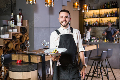 Plakat Przystojny młody facet z brodą ubrany w fartuch stoi w restauracji i trzyma biały talerz z ćmy. W tle, kontuar barowy i wnętrze w stylu loftu