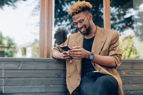 Plakat Portret African American człowieka w kurtkę i okulary z smartphone siedzi w pobliżu okna