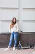 Beautiful young girl with long brown hair stopped while riding the scooter on the background of the gray wall. She is dressed in a white sweater and blue jeans Beautiful young girl with long brown hair stopped while riding the scooter on the background of the gray wall. She is dressed in a white sweater and blue jeans