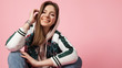 Portrait of a fashionable beautiful laughing girl in a sports jacket print and jeans sitting on a pink background Portrait of a fashionable beautiful laughing girl in a sports jacket print and jeans sitting on a pink background