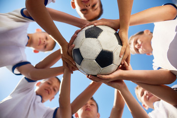 Low angle view of boys in junior football team standing in circle holding ball together against  blue sky, focus on ball