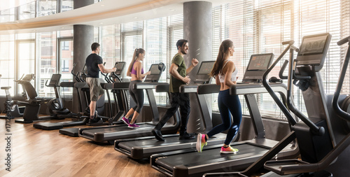 Plakat Grupa czterech osób, mężczyzn i kobiet, biegających na bieżni w nowoczesnej i świetlistej siłowni fitness