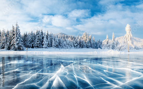 Blue ice and cracks on the surface of the ice. Frozen lake under a blue sky in the winter. The hills of pines. Winter. Carpathian, Ukraine, Europe. © standret