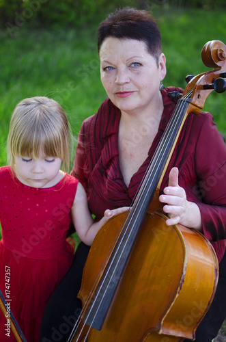 Plakat Stare kobiety i blond caucasian dziewczyna bawić się koncert na wiolonczeli outdoors. Szczęśliwa babcia uczy grać wiolonczela jej wnuczka.