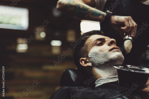 Crop stylish applying foam on customer's cheeks for shaving while working in barbershop. © kkolosov