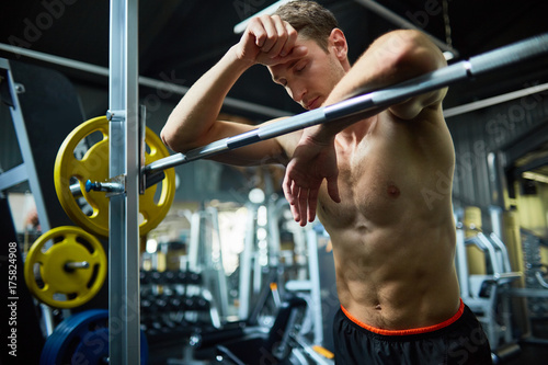 Plakat Talia portret mięśniowy młody człowiek z zamkniętymi oczami opiera na barbell podczas gdy brać przerwę od intensywnego treningu przy przestronnym gym