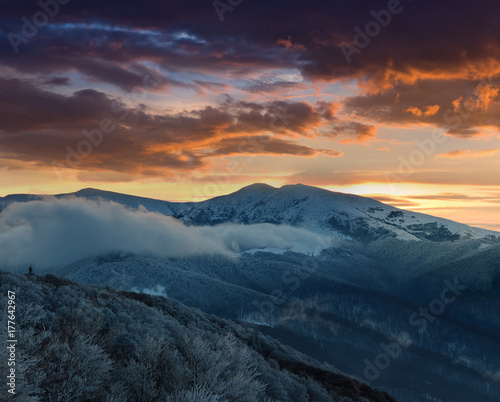 Beautiful sunrise in the winter mountains. Dramatic cloudy over sky. View of foggy hills  and trees covered with rime. 