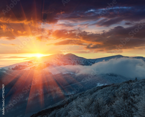 Beautiful sunrise in the winter mountains. Dramatic cloudy over sky. View of foggy hills  and trees covered with rime. 