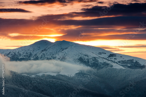 Beautiful sunrise in the winter mountains. Dramatic cloudy over sky. View of foggy hills  and trees covered with rime. 
