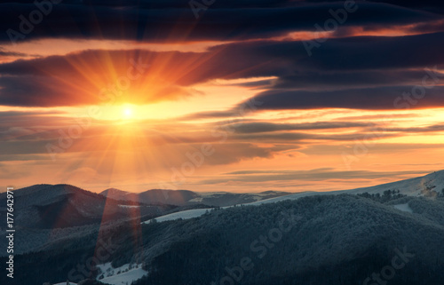 Beautiful sunrise in the winter mountains. Dramatic cloudy over sky. View of foggy hills  and trees covered with rime. 