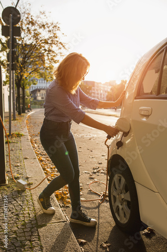 Zdjęcie XXL Młoda kobieta ładuje elektryczny samochód. Wypożyczany samochód ładuje się w stacji ładowania pojazdów elektrycznych. Dzielenie samochodów.