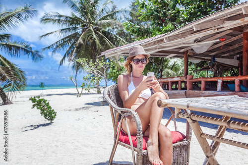 A beautiful young woman in a white bikini sits on a tropical beach with a phone in her hands. Rest, vacation, resort, beautiful life