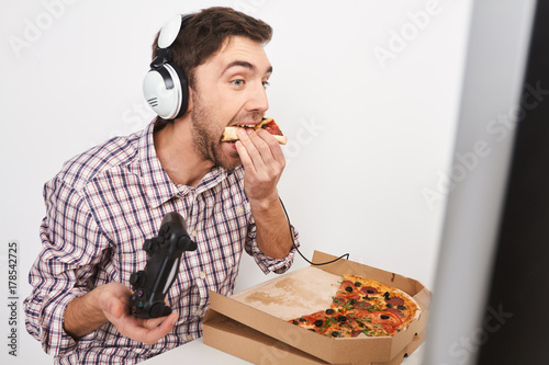 Zdjęcie XXL Niezdrowy tryb życia. Zakończenie młody piękny śmieszny brodaty mężczyzna z ciemnymi krótkimi włosami bawić się online gry z kontrolerem, opowiadający z przyjaciółmi w hełmofonach, je pizzę.