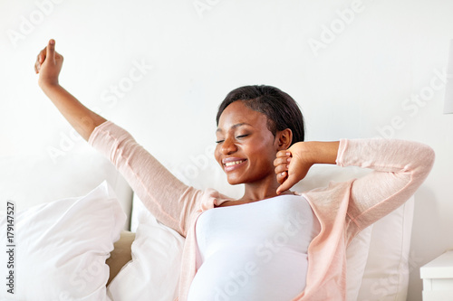 happy pregnant african woman stretching in bed