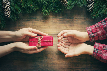 Red gift box in female hands and the child's hands accepting a gift, christmas fir tree on wooden background. Christmas celebration concept.
