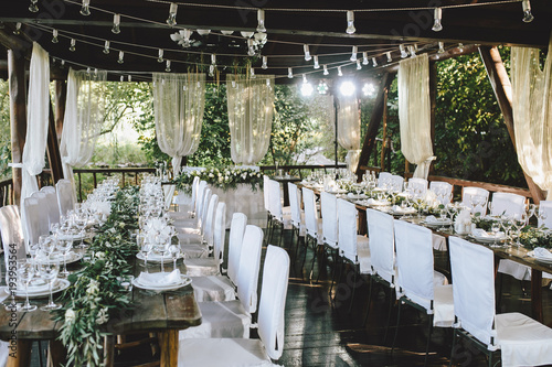 Decorated elegant wooden wedding table for banquet outdoor in garden gazebo with lamp, in the style of rustic with eucalyptus and flowers, porcelain plates, glasses, white chairs