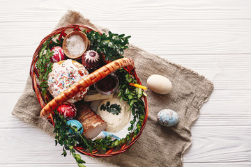 stylish basket with painted eggs, bread, ham,beets, butter on rustic wood background with spring flowers and candle, top view. easter food for blessing in church. happy Easter concept