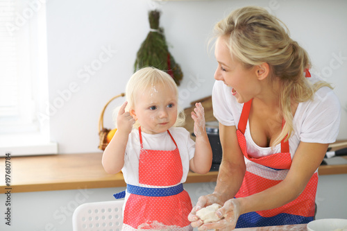 Plakat Mała dziewczynka i jej blondynki mama w czerwonych fartuchach bawić się i śmia się podczas gdy ugniatający ciasto w kuchni. Domowe ciasta na chleb, pizzę lub upiecz ciasteczka