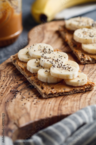 Plakat Dwa wieloziarniste pieczywo z pastą z orzeszków ziemnych, plasterkami banana i nasionami chia na drewnianej desce do serwowania. Koncepcja zdrowego śniadania fitness.