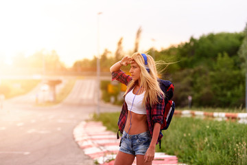 Beautiful young woman traveler with backpack walking along highway in sunset