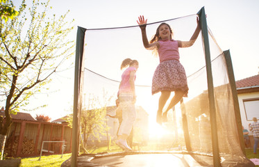 Three charming cute kids are jumping on the trampoline and the youngest girl is attempting to jump over the safety net during a beautiful sunny day.