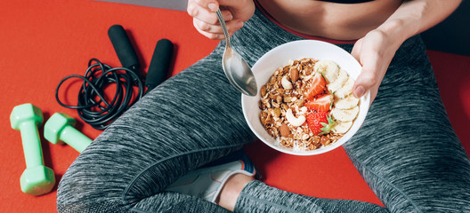 girl in sports clothes sitting near dumbbells and skipping rope with a plate of healthy breakfast in her hands