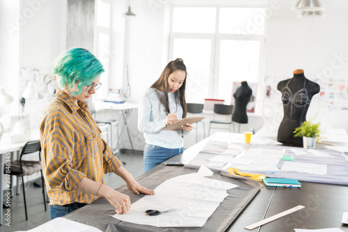 Фото: Wide angle portrait of two women standing at tailors table and working in modern design atelier, copy space Wide angle portrait of two women standing at tailors table and working in modern design atelier, copy space