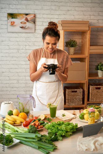 Фото: Attractive middle-aged food blogger wearing apron taking picture of healthy smoothie while standing at modern kitchen Attractive middle-aged food blogger wearing apron taking picture of healthy smoothie while standing at modern kitchen