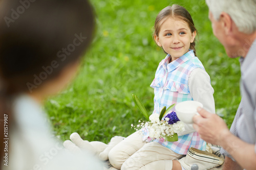 Фото: Cute little girl with floral bouquet looking at grandfather during talk by cup of tea on picnic Cute little girl with floral bouquet looking at grandfather during talk by cup of tea on picnic