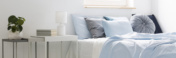 Real photo of a bed with blue bedding and cushions standing next to white tables with books, lamp and plant in bedroom interior