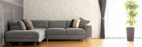 Фото: Grey corner sofa with decorative cushions in the real photo of bright living room interior with brick wall, fresh green plant and window with drapes Grey corner sofa with decorative cushions in the real photo of bright living room interior with brick wall, fresh green plant and window with drapes