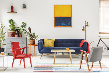 Red armchair next to wooden table and blue sofa in living room interior with painting. Real photo