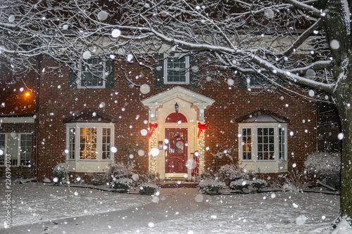 Snowfall on beautiful brick house with columns and bay windows with Christmas tree light up and red sled and wreath on porch © Susan Vineyard 