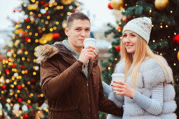 christmas portrait of happy couple with hot mulled wine or tea walking on city streets decorated for holidays, christmas trees with lights and toys on background