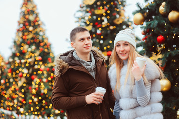 christmas portrait of happy couple with hot mulled wine or tea walking on city streets decorated for holidays, christmas trees with lights and toys on background
