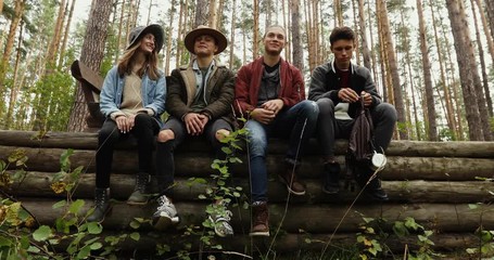 Four young people in jeans and jackets sitting on pile of logs in forest on background of trees. One guy is sad he is offended