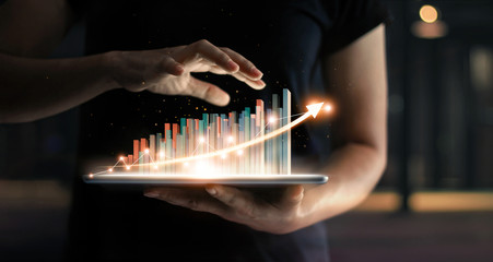 Businessman holding tablet and showing a growing virtual hologram of statistics, graph and chart with arrow up on dark background. Stock market. Business growth, planing and strategy concept.