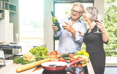 Senior couple cooking healthy food and drinking red wine at house kitchen - Retired people at home preparing lunch with fresh vegetables and bio products - Happy elderly concept with mature pensioner