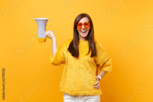 Portrait of cheerful attractive young woman in fur sweater orange heart eyeglasses holding megaphone isolated on bright yellow background. People sincere emotions, lifestyle concept. Advertising area. © ViDi Studio